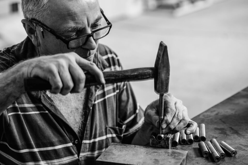 Artigianatoprime | Trova i migliori artigiani in tutta Italia An elderly craftsman skillfully hammers metal in a workshop, showcasing artisanal skill and precision.